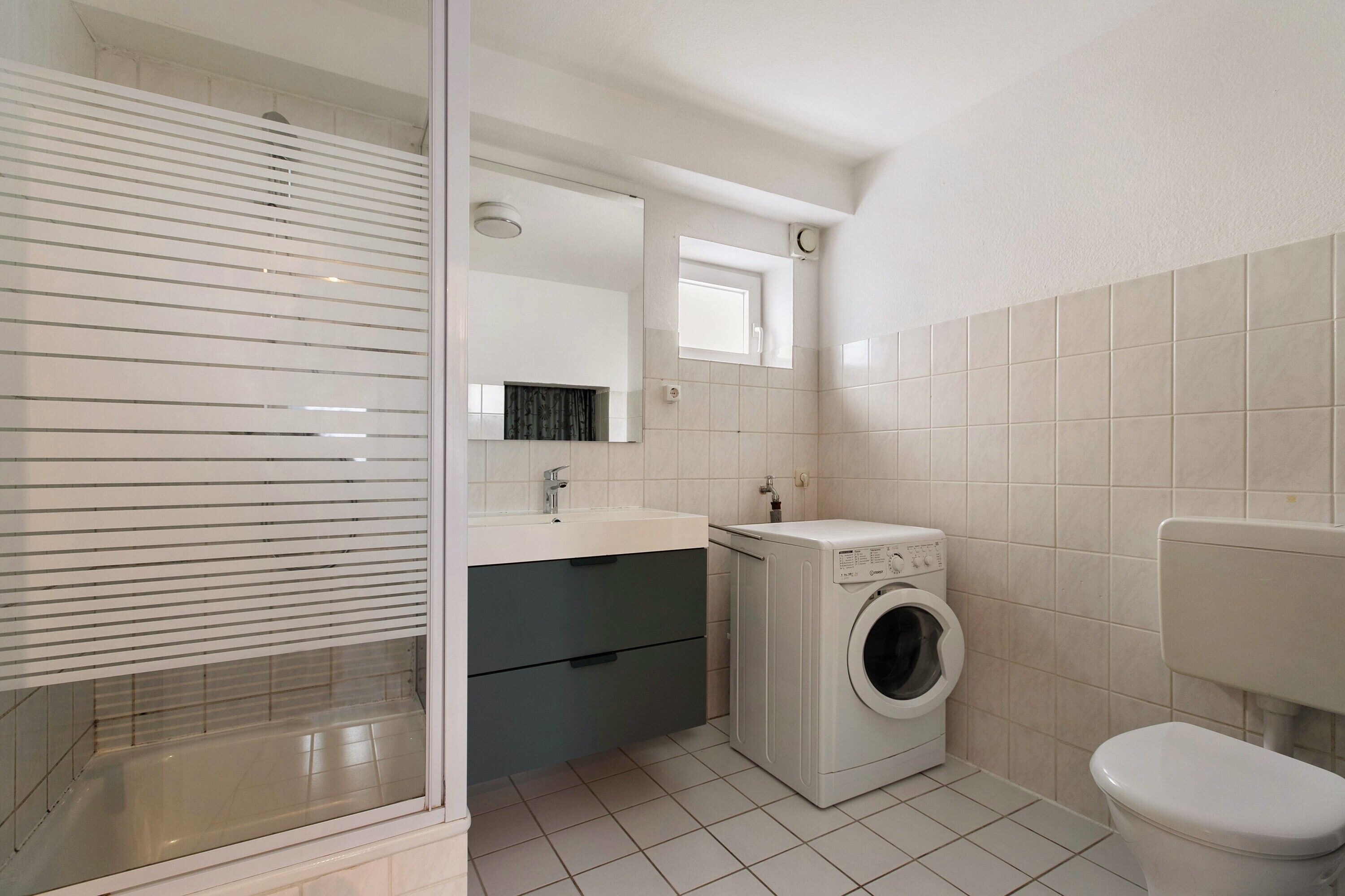 A compact bathroom with a washing machine, a toilet, a sink with a mirror, and a window with blinds.