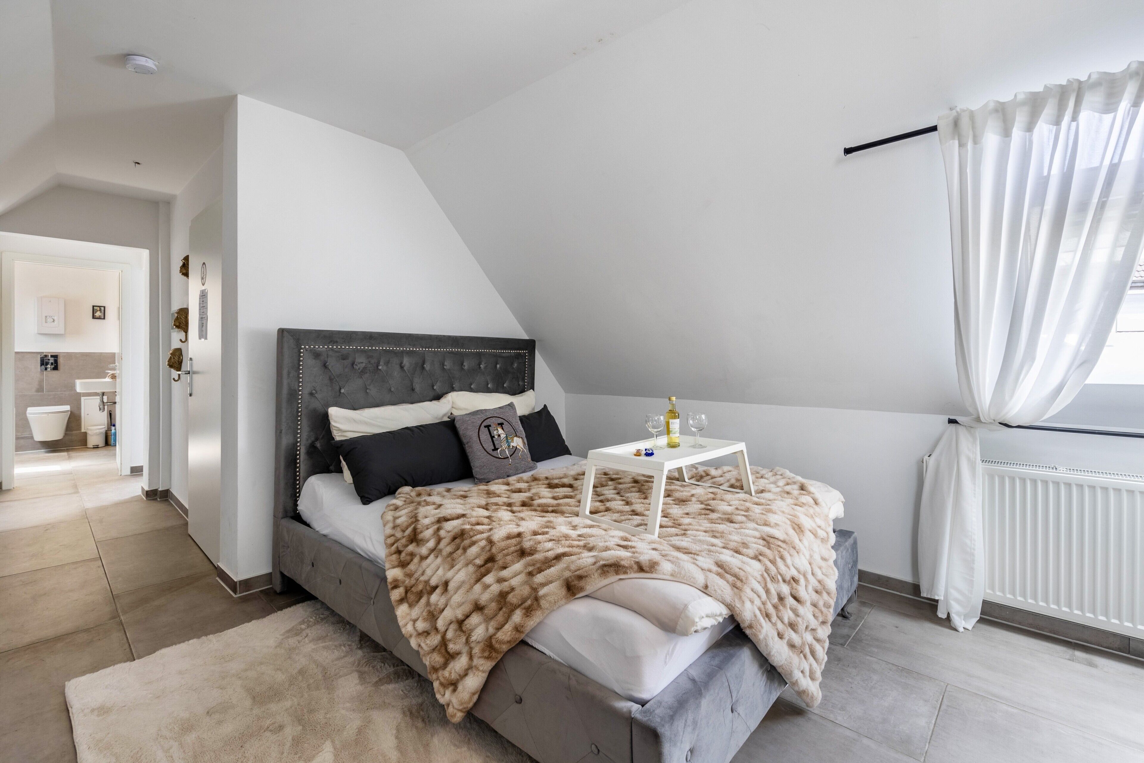 Ein Schlafzimmer mit einem Bett, einem Nachttisch mit Lampe, einem Fenster mit Vorhängen und Blick ins Bad.