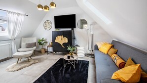 A modern loft bedroom with a black sofa, a white armchair, a round coffee table, and a large window with white curtains. - Apartment 'Deluxe' with Wi-Fi and Air Conditioning (Duisburg)