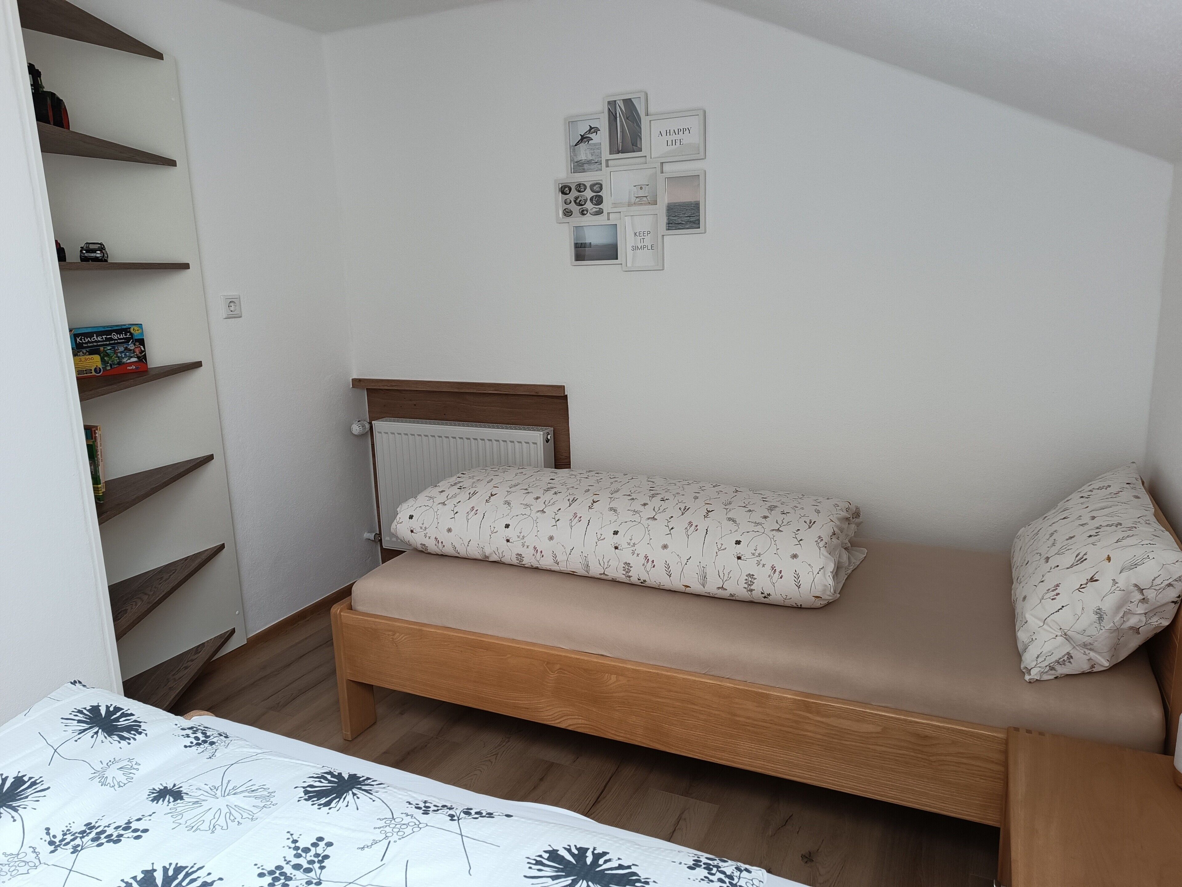 Quarto pequeno com uma cama de solteiro, cabeceira de madeira, radiador, prateleira com livros e parede com quadros.