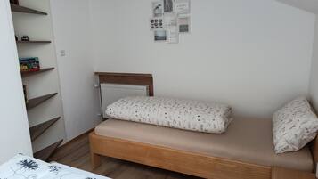 Quarto pequeno com uma cama de solteiro, cabeceira de madeira, radiador, prateleira com livros e parede com quadros.