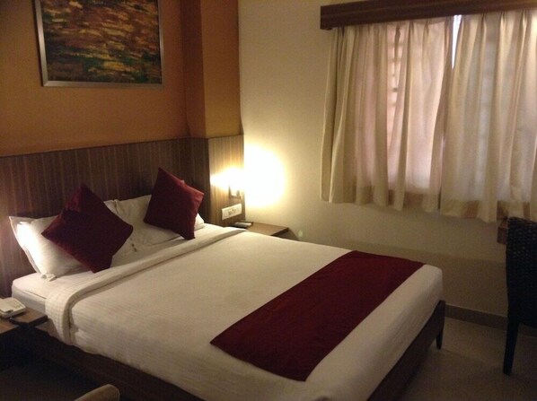 A hotel room with a large bed, a nightstand with a lamp, a painting on the wall, and a window with curtains. - La Sara Comforts (Bengaluru)