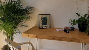 Ein Essbereich mit einem Holztisch, einem weißen Stuhl mit Sitzkissen mit Blattmuster, einer Topfpflanze und einem gerahmten Bild an der Wand.
