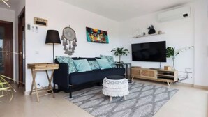 Ein modernes Wohnzimmer mit einem dunkelblauen Sofa, einem kleinen weißen Hocker, einem gemusterten Teppich, einem Flachbildfernseher und einem Traumfänger an der Wand.