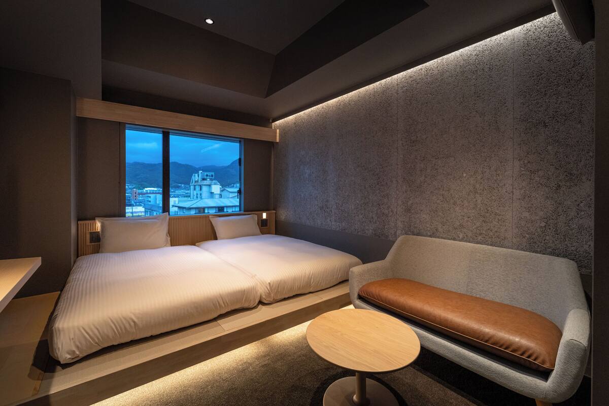 Una habitación de hotel moderna con una cama grande, un sofá y una mesita. Hay una ventana con vistas a la ciudad.