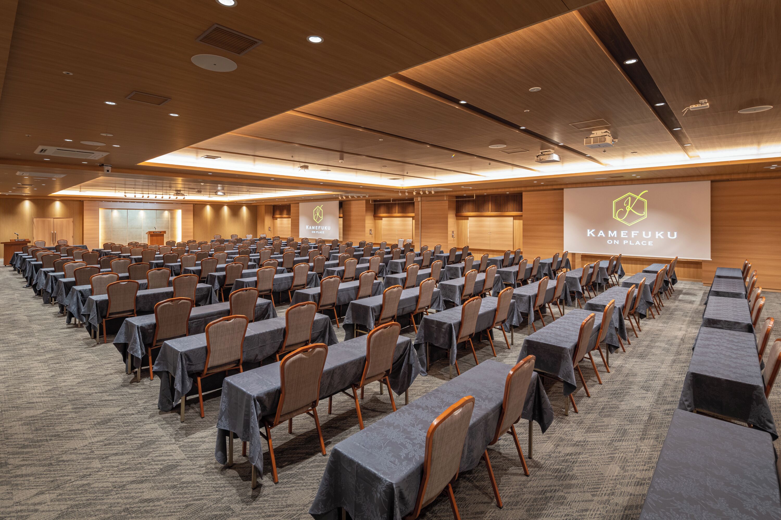 Un salón de conferencias con filas de mesas y sillas, un atril y una pantalla para presentaciones con el logo "KAMEFUKU ON PLACE".