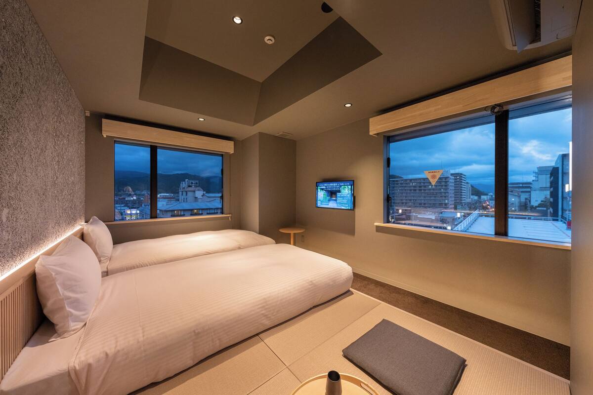 Una habitación de hotel moderna con dos camas, ventanas amplias con vistas a la ciudad, una mesita y un televisor en la pared.