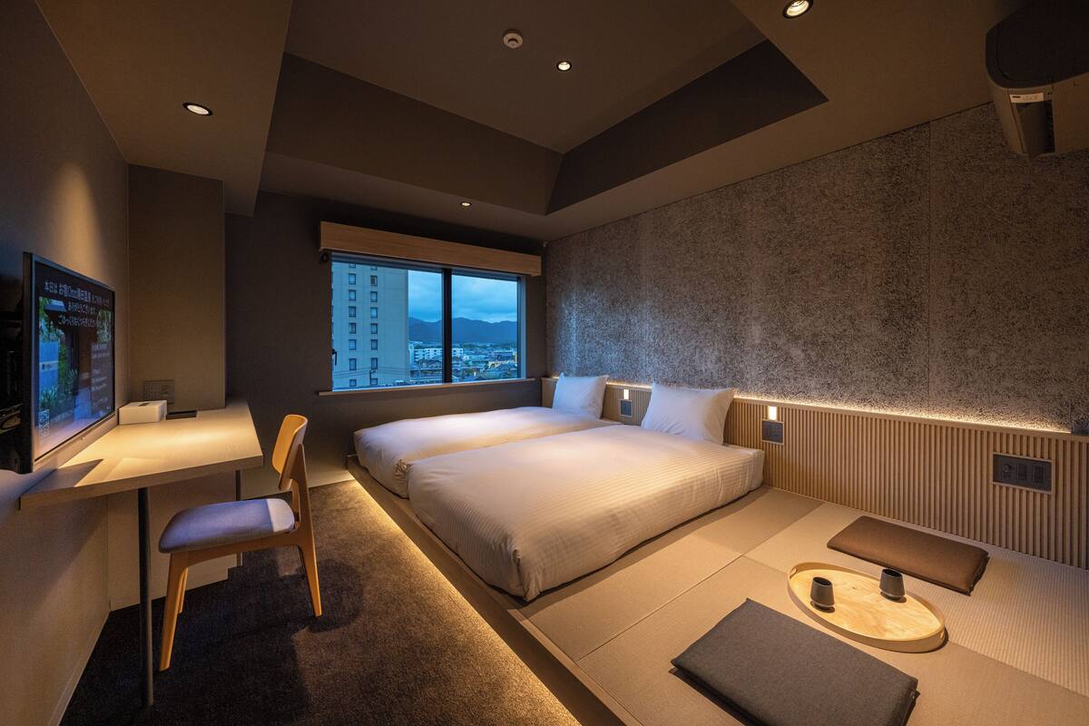Habitación de hotel moderna con una cama grande, un escritorio y una silla. Se ve una ventana con vista a la ciudad.