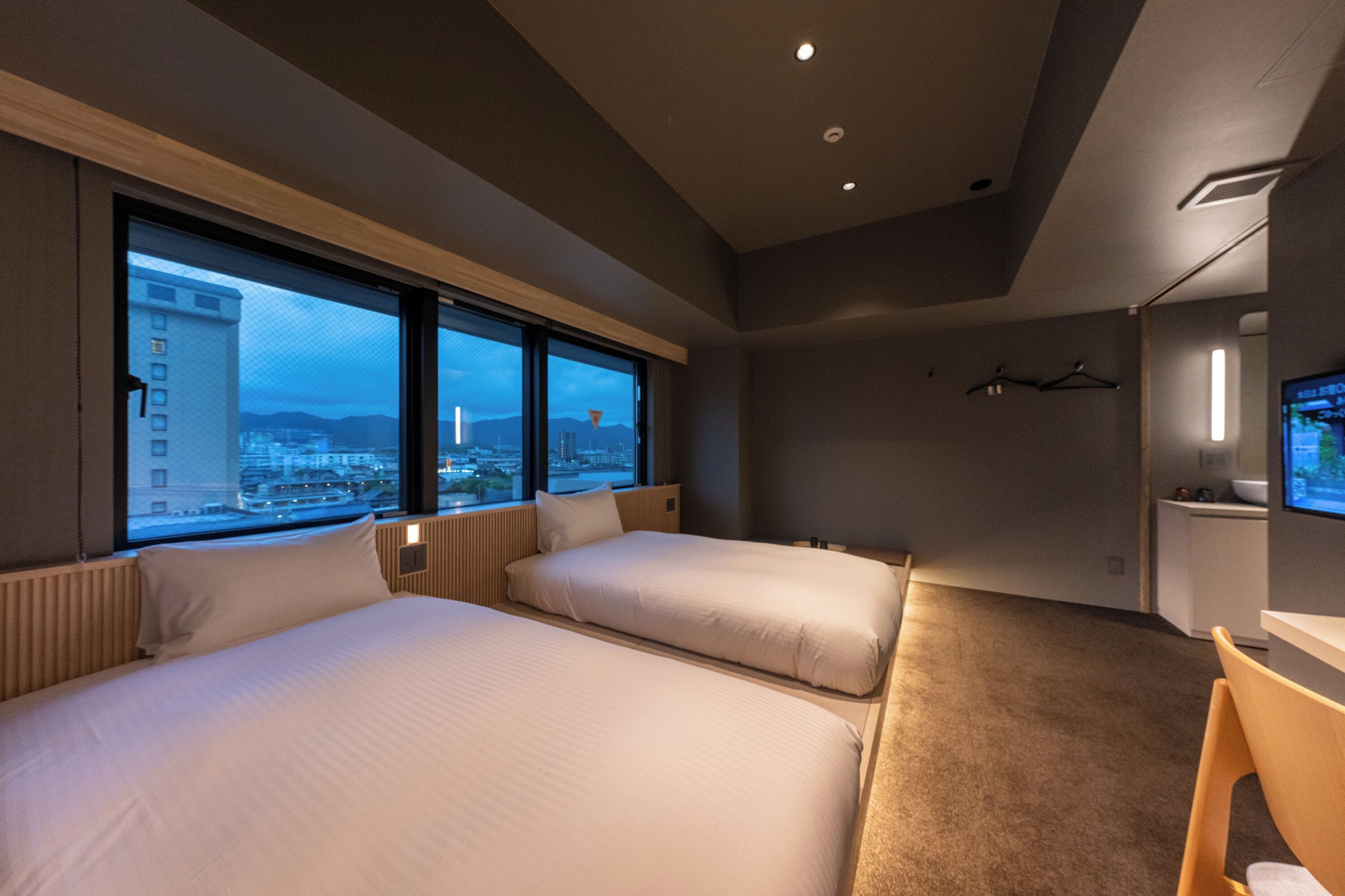 A hotel room with two beds, a desk, a TV, and a large window offering a city view.