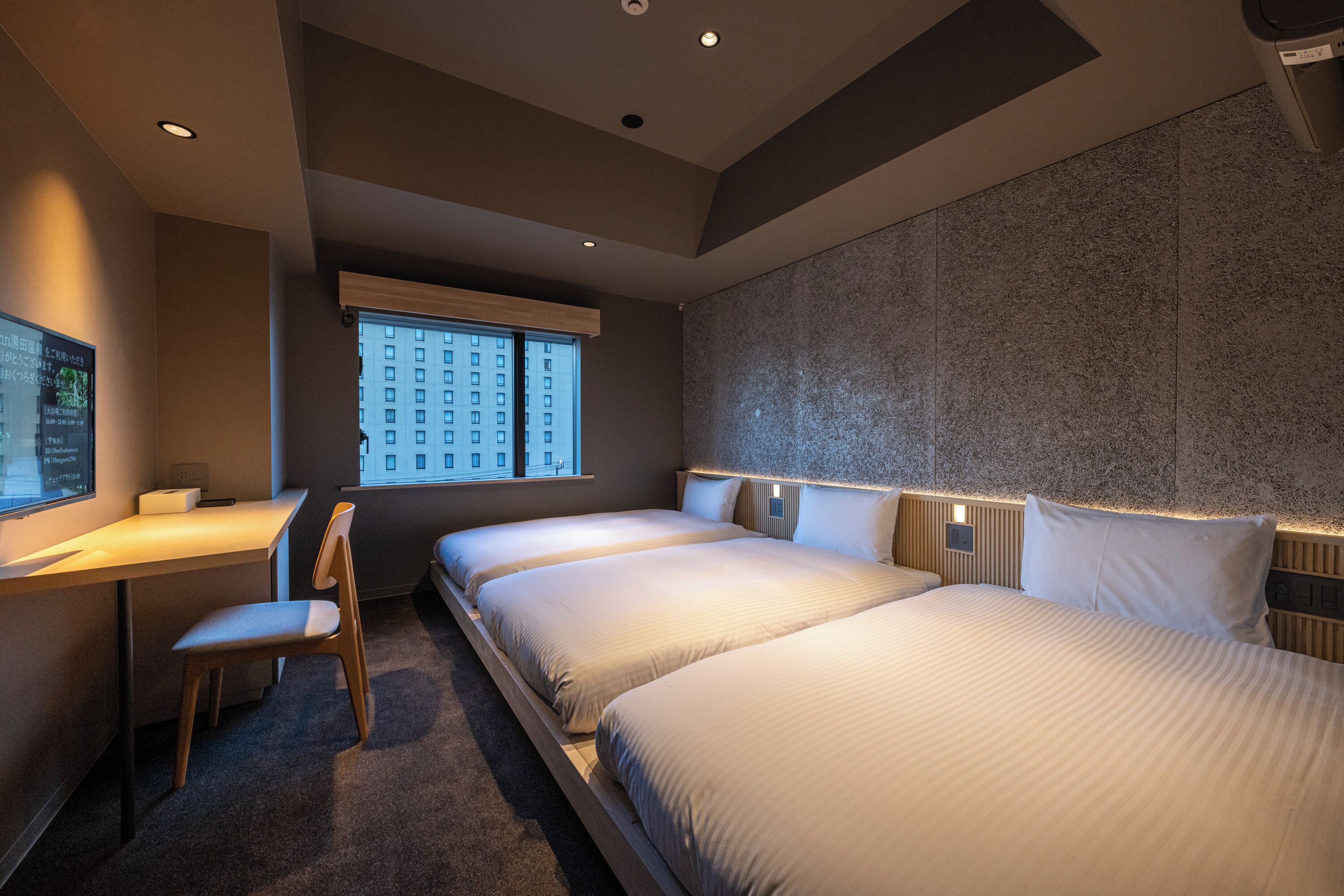 A hotel room with a desk, two beds, a chair, and a large window with a city view.