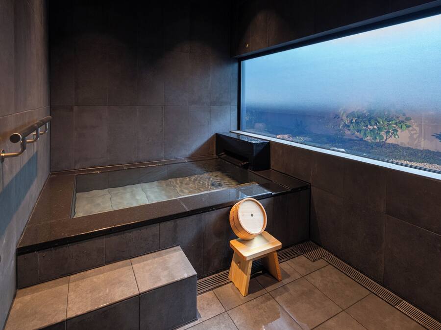 A modern bathroom with a large bathtub, a wooden stool, and a window offering a view of the outdoors.
