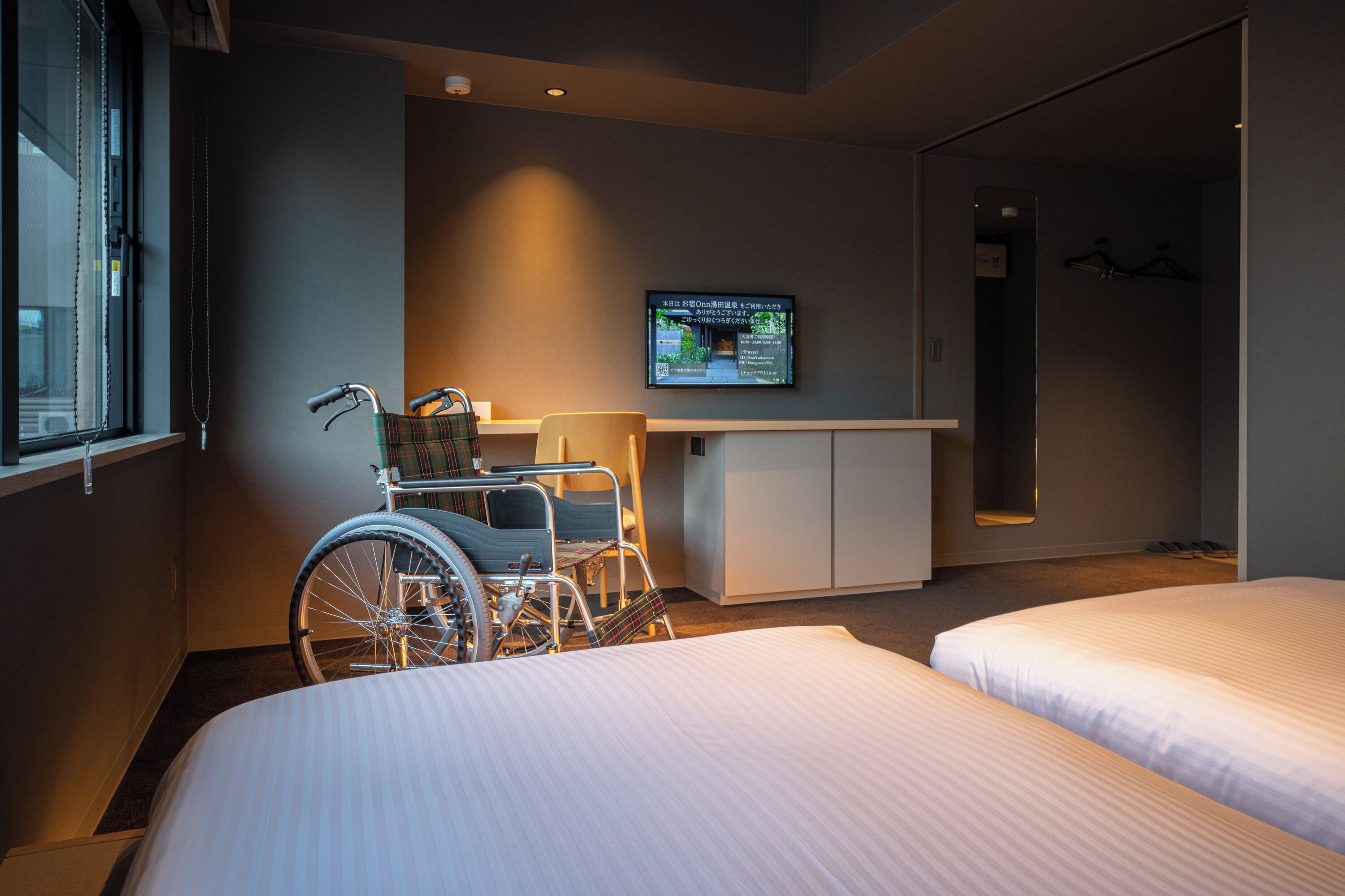 A hotel room with two beds, a wheelchair, a desk, and a television mounted on the wall.