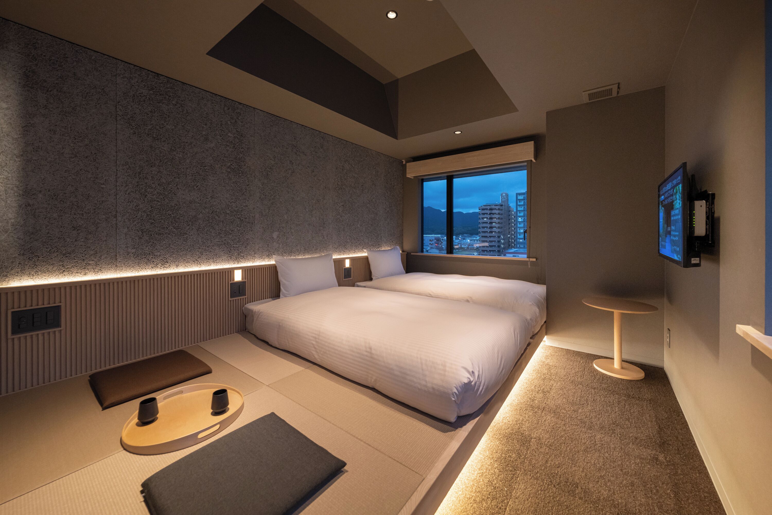 A modern bedroom with a large bed, bedside tables, a small round table, and a view of the city at dusk.