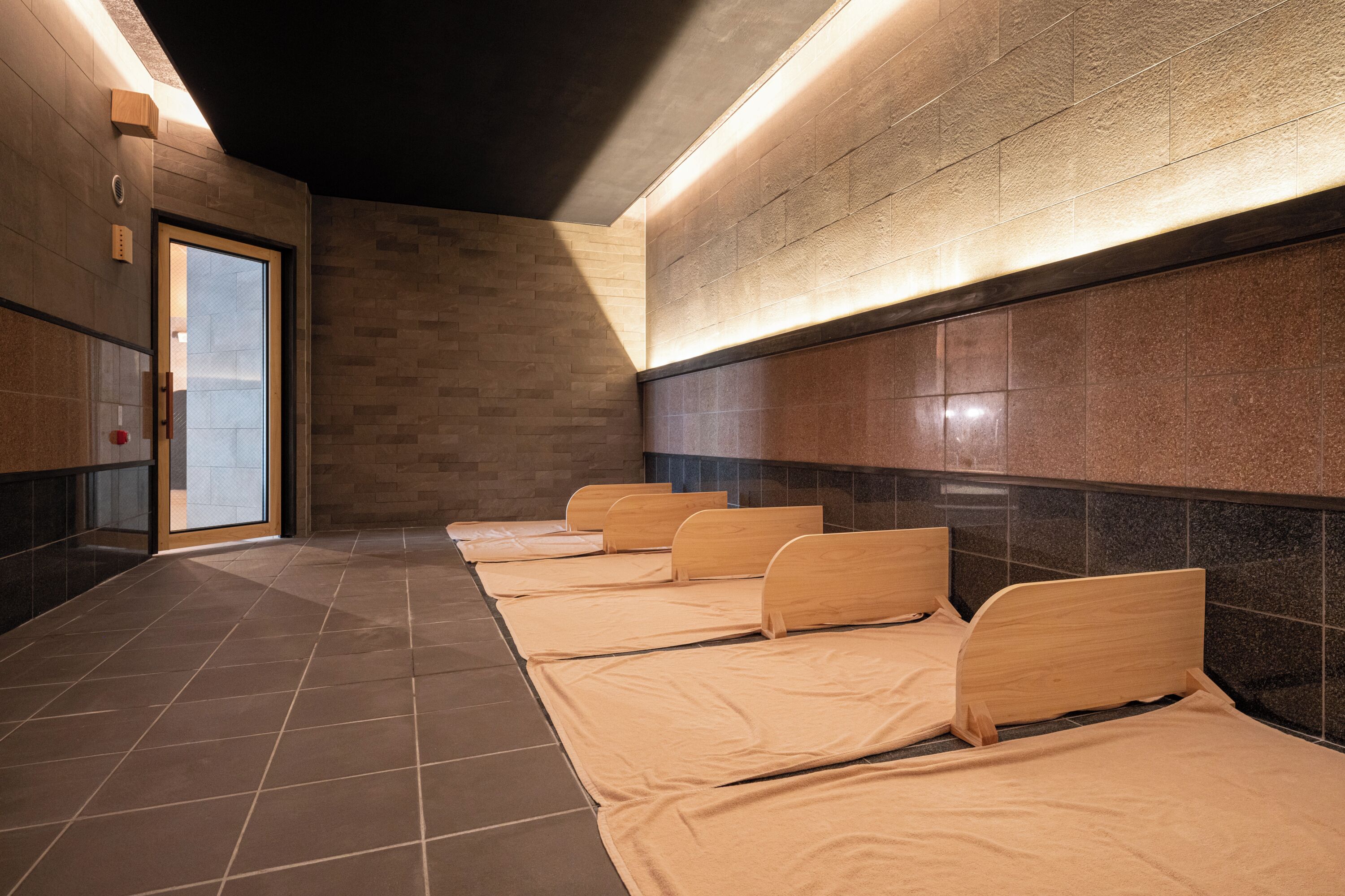 A row of wooden beds in a hallway with tiled floors and walls, a door on the left, and a wall-mounted light fixture.