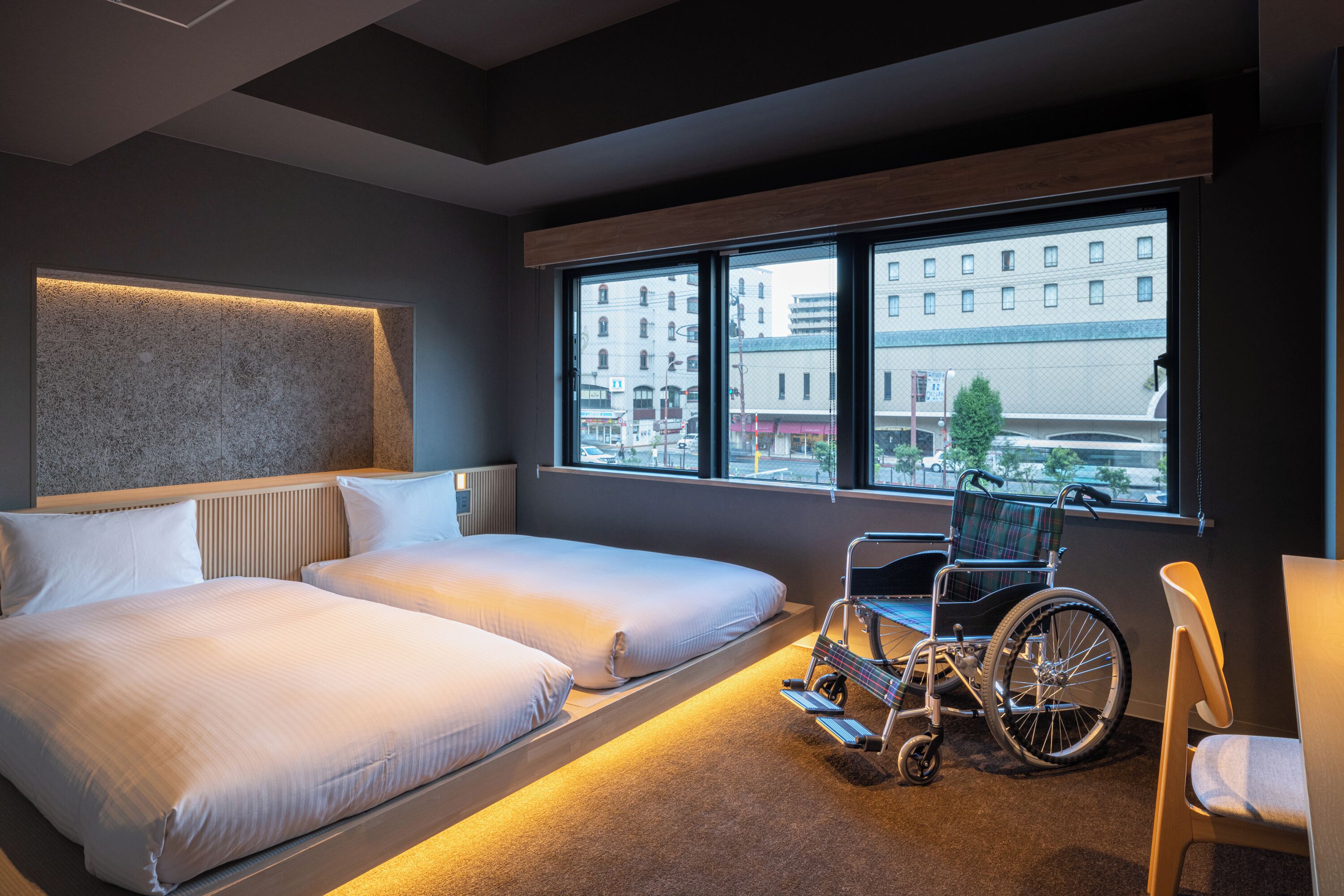 A hotel room with two beds, a wheelchair, and a view of the city through large windows.