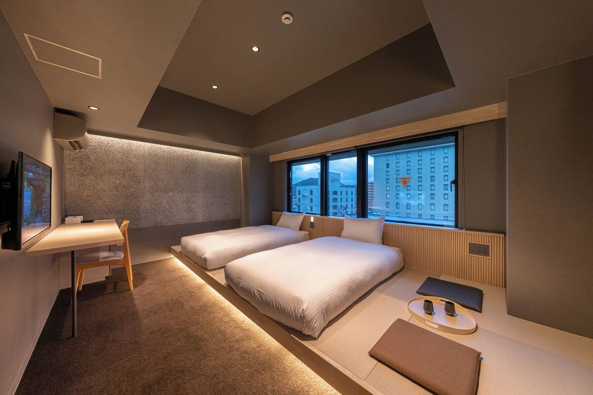 Una habitación de hotel moderna con escritorio, dos camas, un ventanal grande con vista a la ciudad y un televisor de pared.