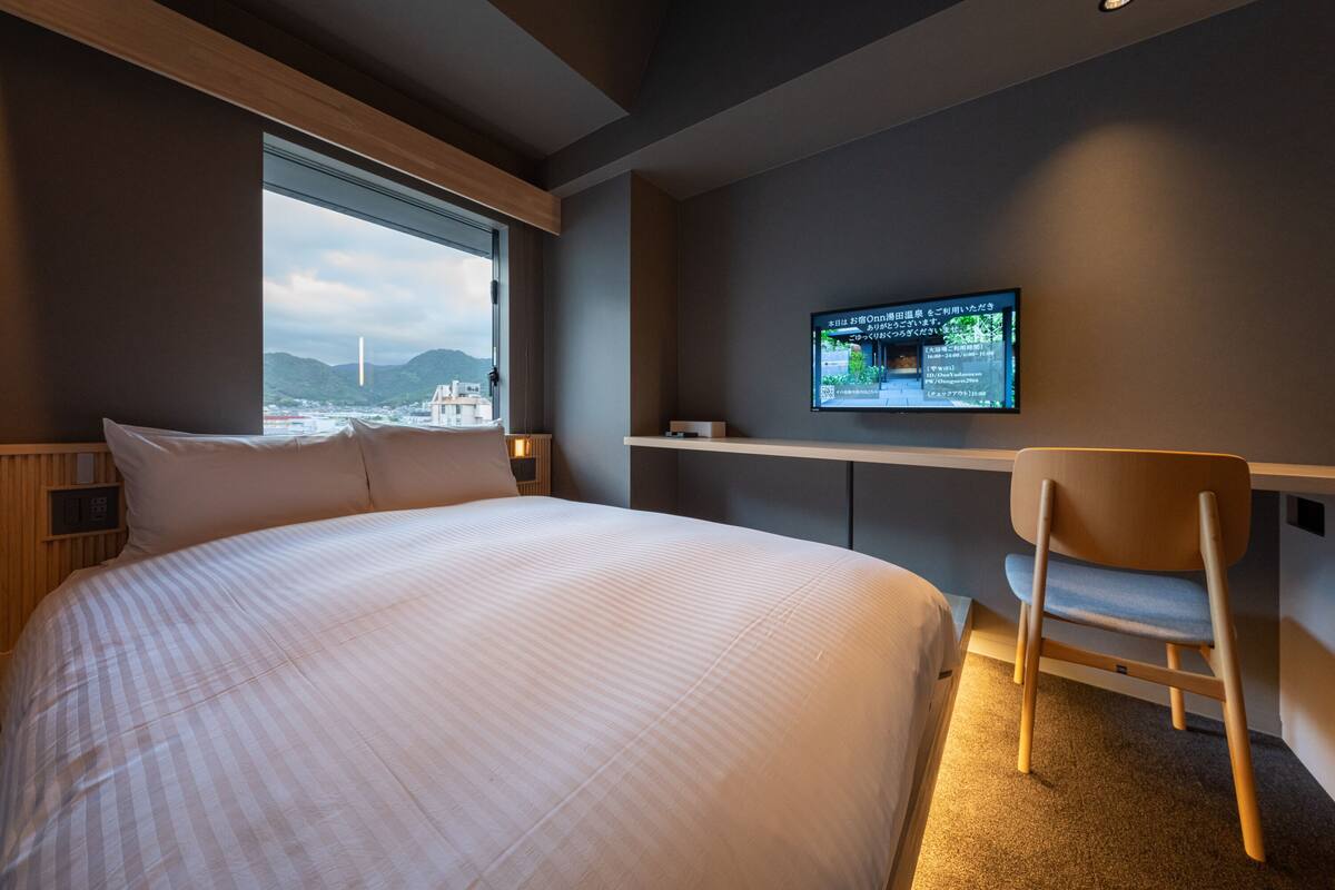 Una habitación de hotel moderna con una cama grande, un televisor montado en la pared y vistas a la ciudad a través de un ventanal amplio.