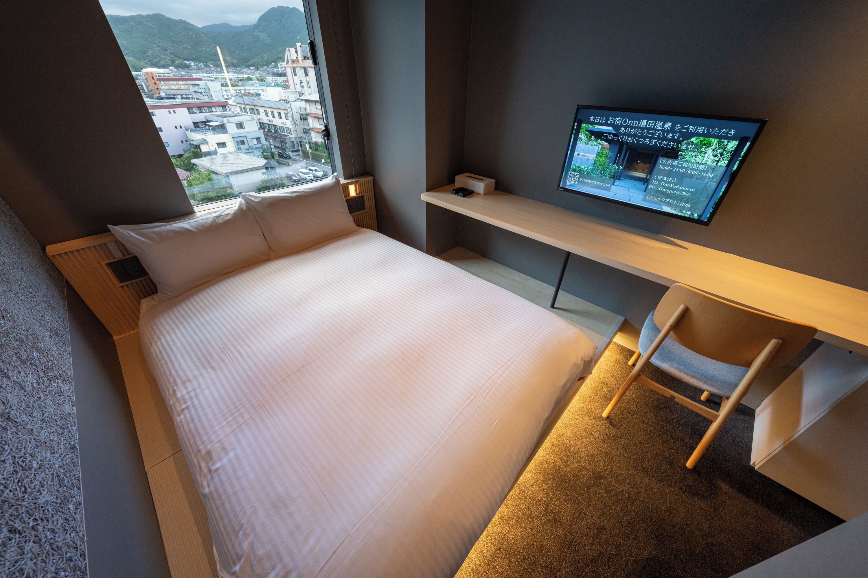 A hotel room with a bed, a desk, a television, and a view of buildings through a window.