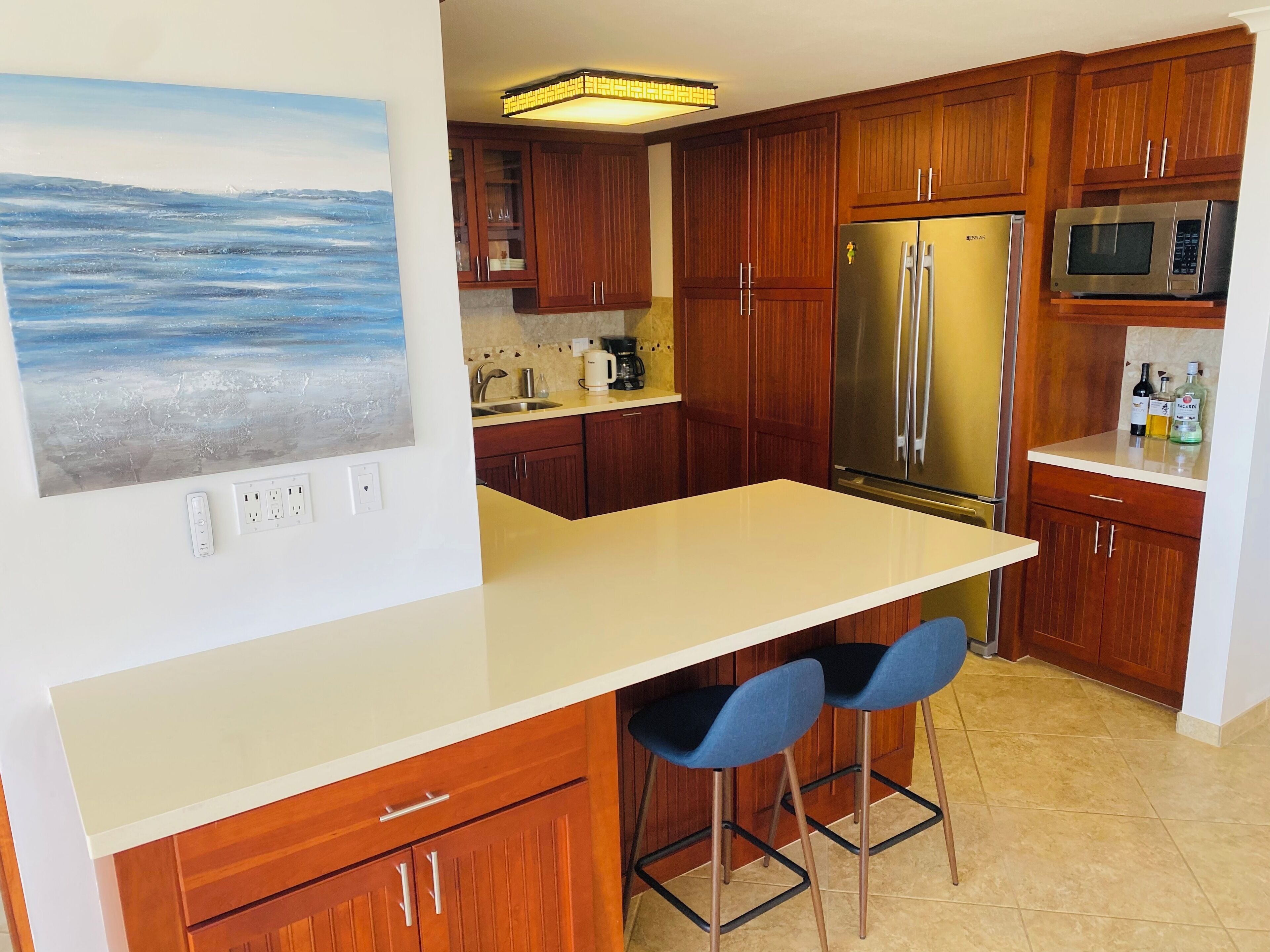 A modern kitchen with wooden cabinets, a stainless steel refrigerator, a microwave, and a bar area with two stools.
