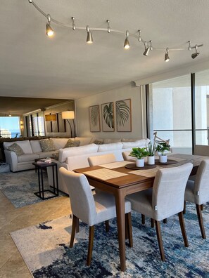 A modern living room with a dining area, featuring a sofa set, a wooden dining table with chairs, and a string of pendant lights.