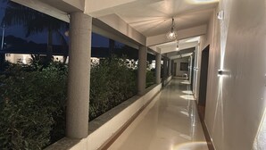 Un pasillo con suelo de baldosas, paredes blancas y columnas. Hay plantas a la izquierda y una lámpara colgante en el centro.