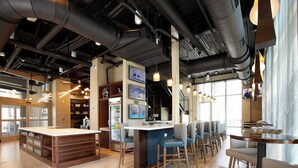 A modern bar area with a long counter, multiple seating options, and visible kitchen equipment in the background. - Holiday Inn Louisville Downtown (Louisville)