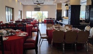 A dining area with round tables and chairs, a kitchen in the background, and large windows. - Rock Royal Hotel and Resort (Kep)