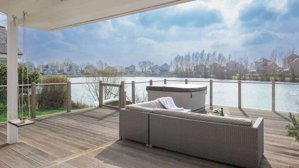 Una terraza cubierta al aire libre con un sofá de mimbre y un jacuzzi, con vistas a un lago y casas al fondo.