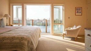 Un dormitorio con una cama grande, una silla, un aparador y vistas a un cuerpo de agua a través de puertas francesas abiertas.