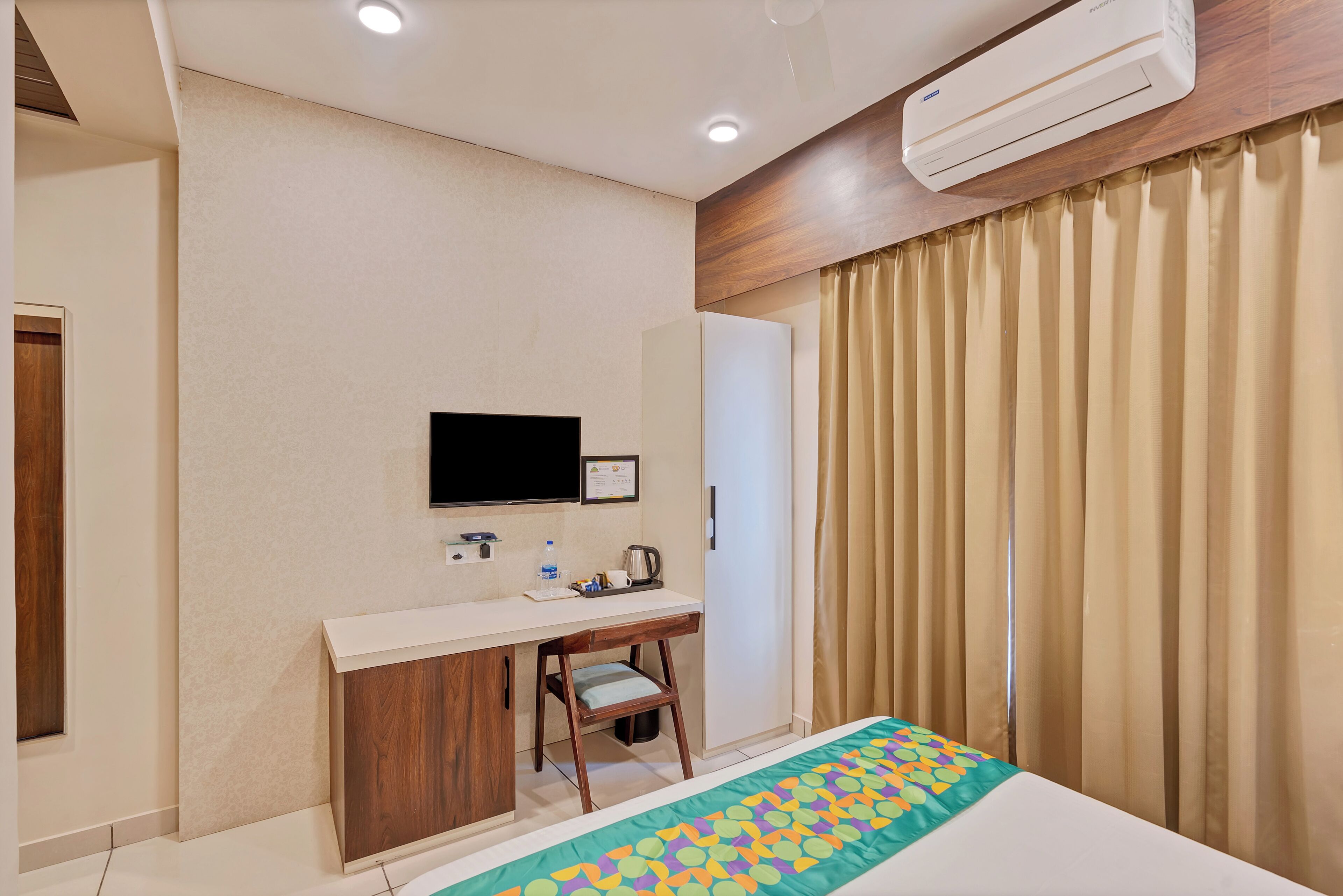A hotel room with a bed, a desk, a chair, a television, an air conditioner, and a window with curtains.