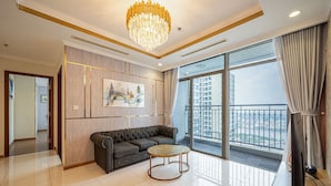 A modern living room with a grey sectional sofa, a round coffee table, a chandelier, and a large window with a city view. - Landmark Condotel Art By KaySuite  (Ho Chi Minh City)