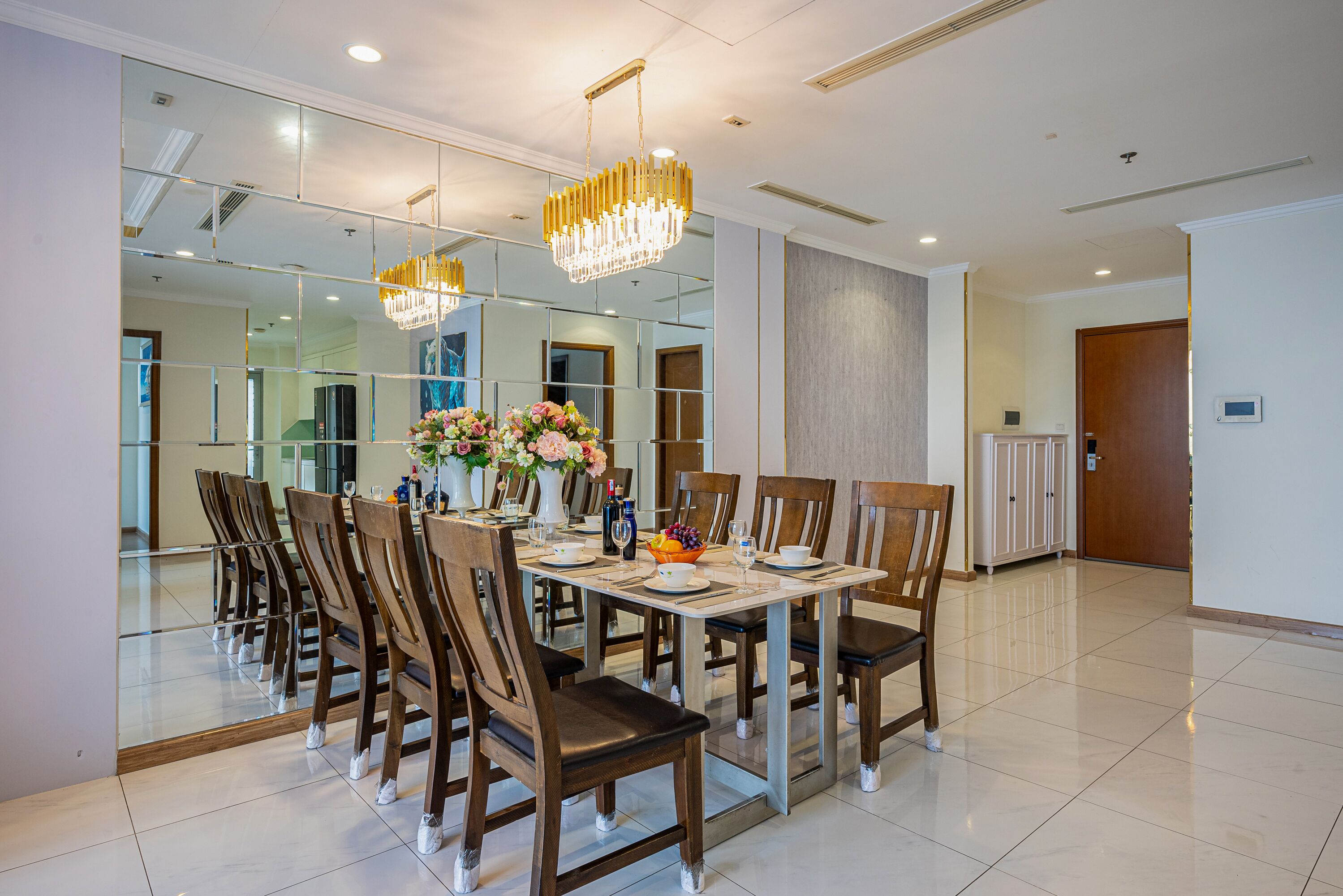 Un comedor con una mesa grande dispuesta para seis personas, rodeada de sillas, con una araña colgante arriba y un espejo que refleja la habitación.