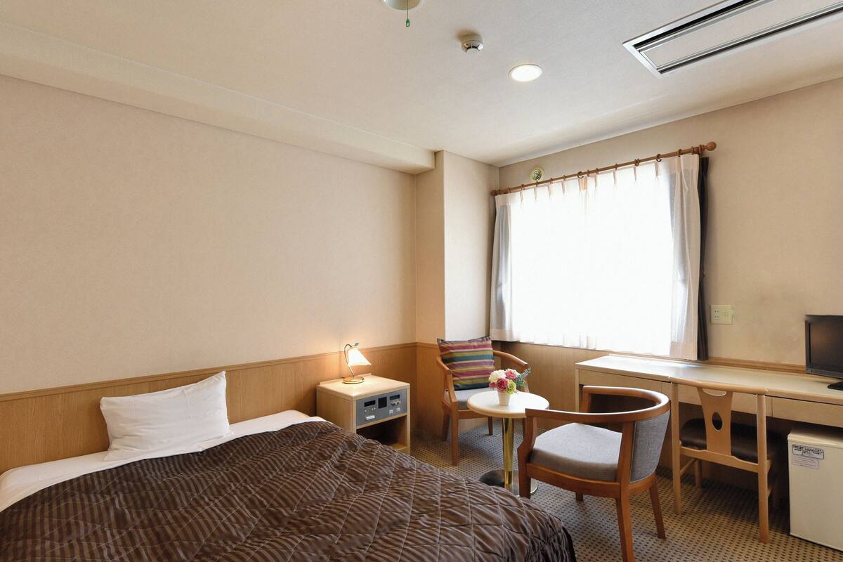 A hotel room with a bed, a desk, a chair, a small table with a lamp, and a television.