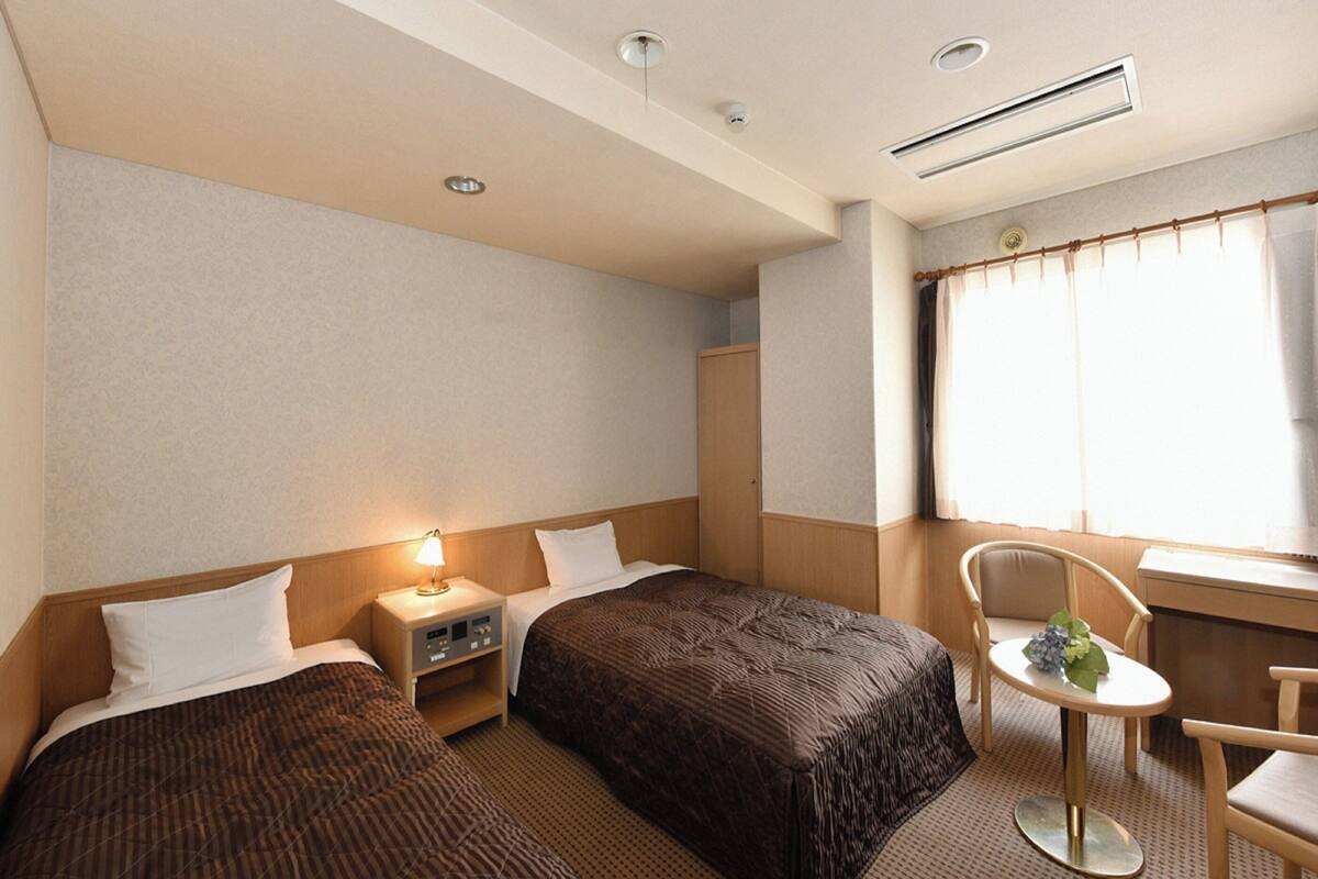 A hotel room with two single beds, a small table with a chair, a nightstand, a window with curtains, and a ceiling vent.