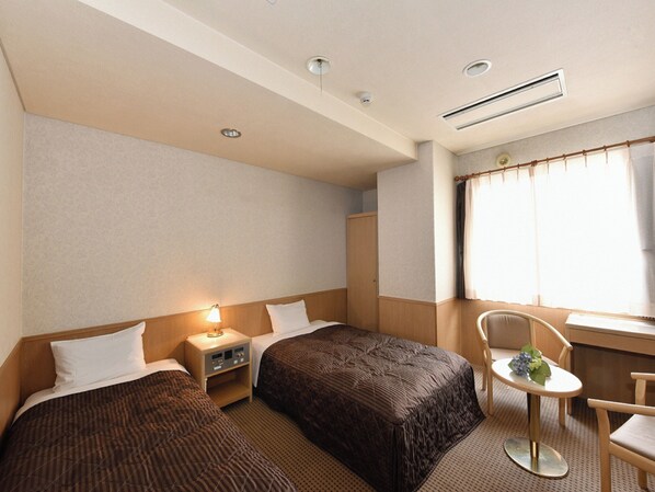 A hotel room with two single beds, a small table with a chair, a nightstand, a window with curtains, and a ceiling vent.