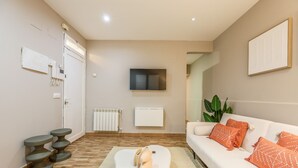 Un salon moderne avec un canapé blanc, des coussins orange, un téléviseur à écran plat, un radiateur et un plancher en bois.