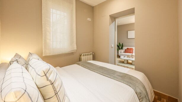 Une chambre à coucher avec un lit, une fenêtre avec des rideaux, un radiateur et une salle de bain visible à travers une porte ouverte.