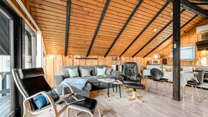A cozy cabin interior with a wooden ceiling, a sofa, a dining table, and a kitchen area.