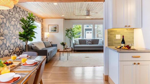 A modern kitchen with white cabinets, a dining area with a table set for breakfast, and a living room with a grey sofa and a plant. - Cove Beach Retreat • Fire Pit + Golf Course Nearby | Grey Oak (Seaside)