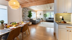 A spacious living area with a dining table set for breakfast, a sofa, and a kitchen with a wine bottle. - Cove Beach Retreat • Fire Pit + Golf Course Nearby | Grey Oak (Seaside)