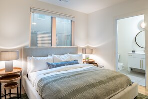 A bedroom with a bed, bedside tables, a window, and a bathroom visible through an open door.