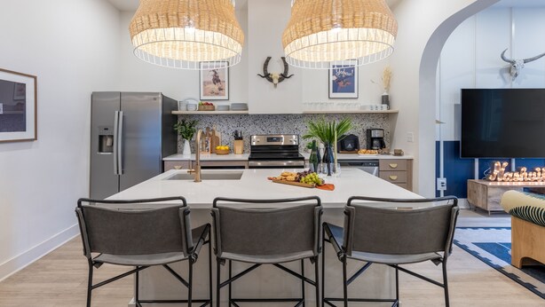 Una cocina moderna con una isla central, dos lámparas colgantes, un refrigerador, una estufa y un televisor en la zona de estar contigua.