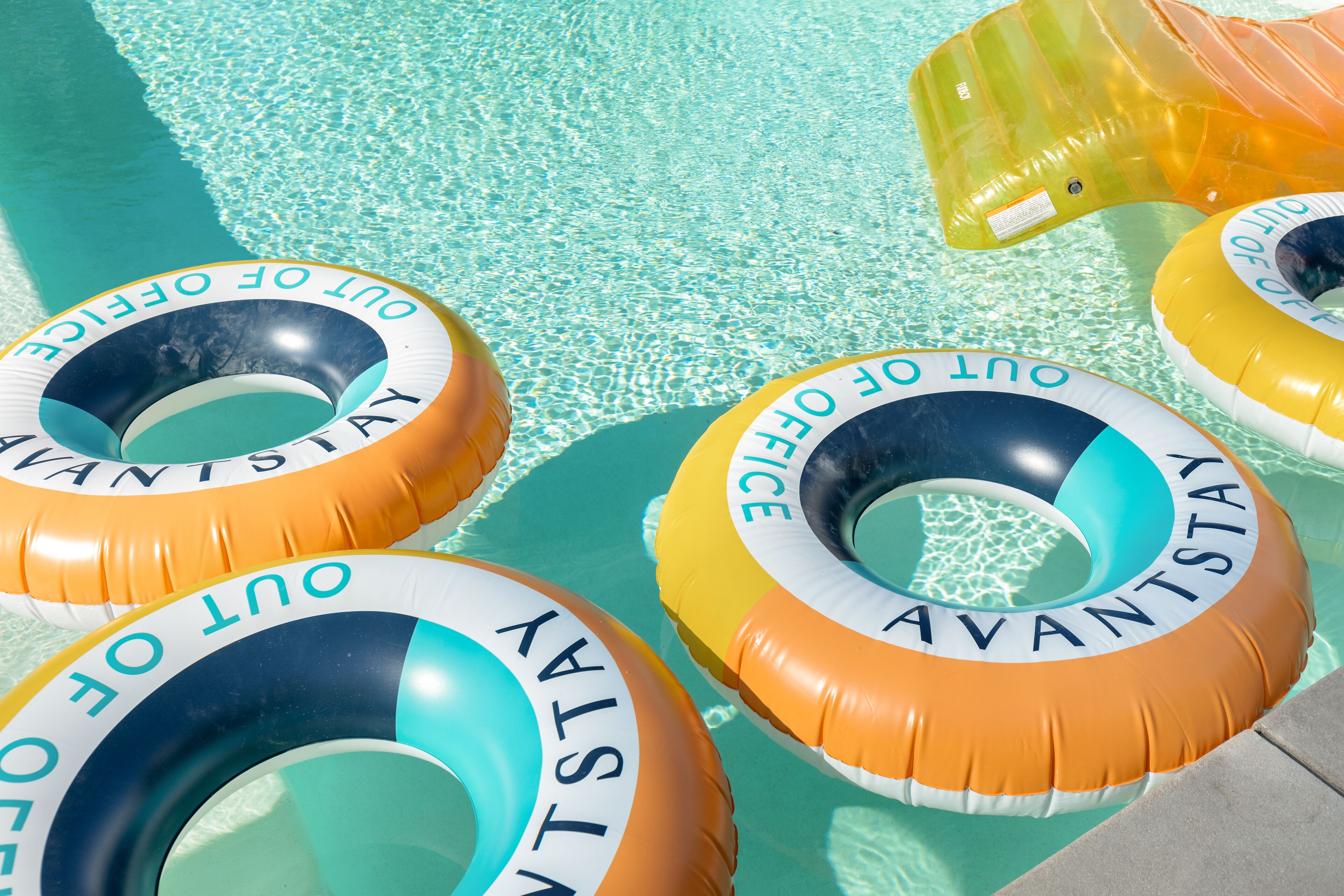 Ein Pool mit schwimmenden Ringen, auf denen 'OUT OF THE WAY' steht, und einem gelben, aufblasbaren Poolnudel.