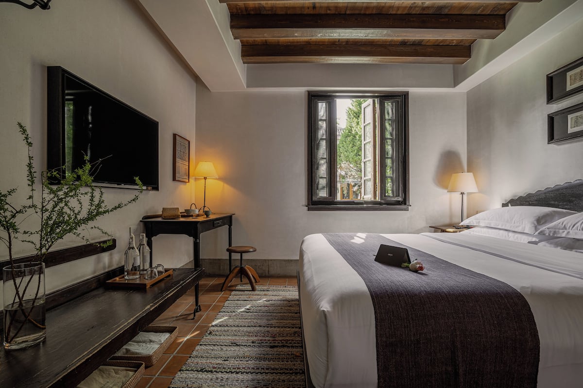 A bedroom with a bed, bedside table, lamp, window, and a TV mounted on the wall.