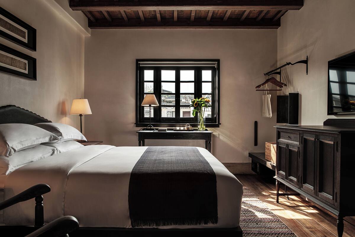 A neatly arranged bedroom with a bed, bedside table, lamp, window, and a dresser.