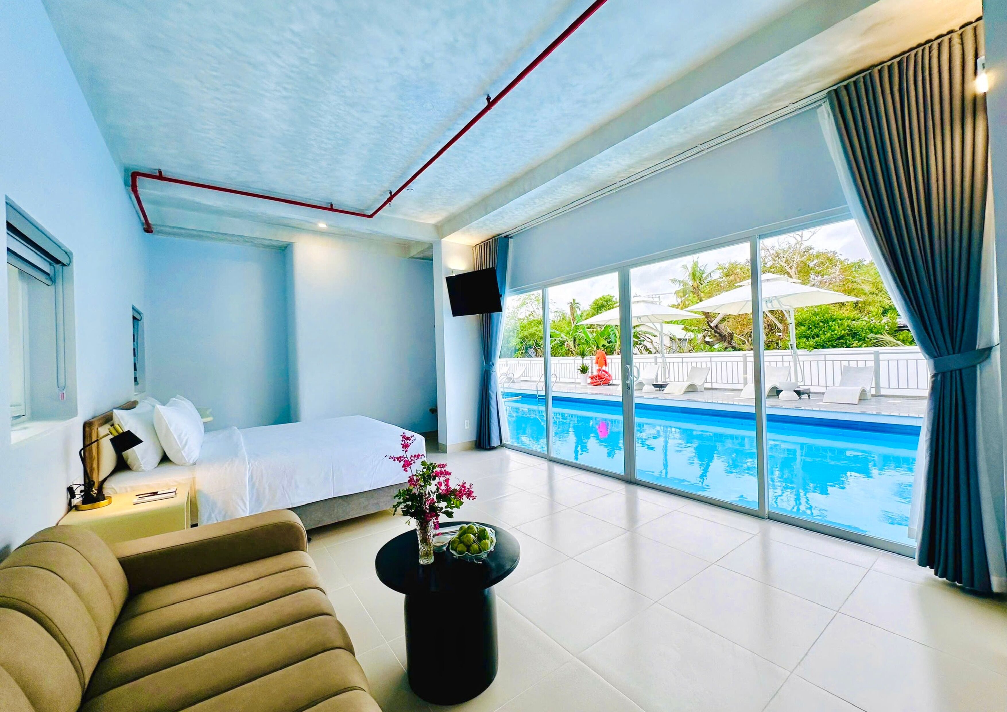 A hotel room with a bed, a sofa, a small table with a vase, a large window with a view of a swimming pool and outdoor seating.