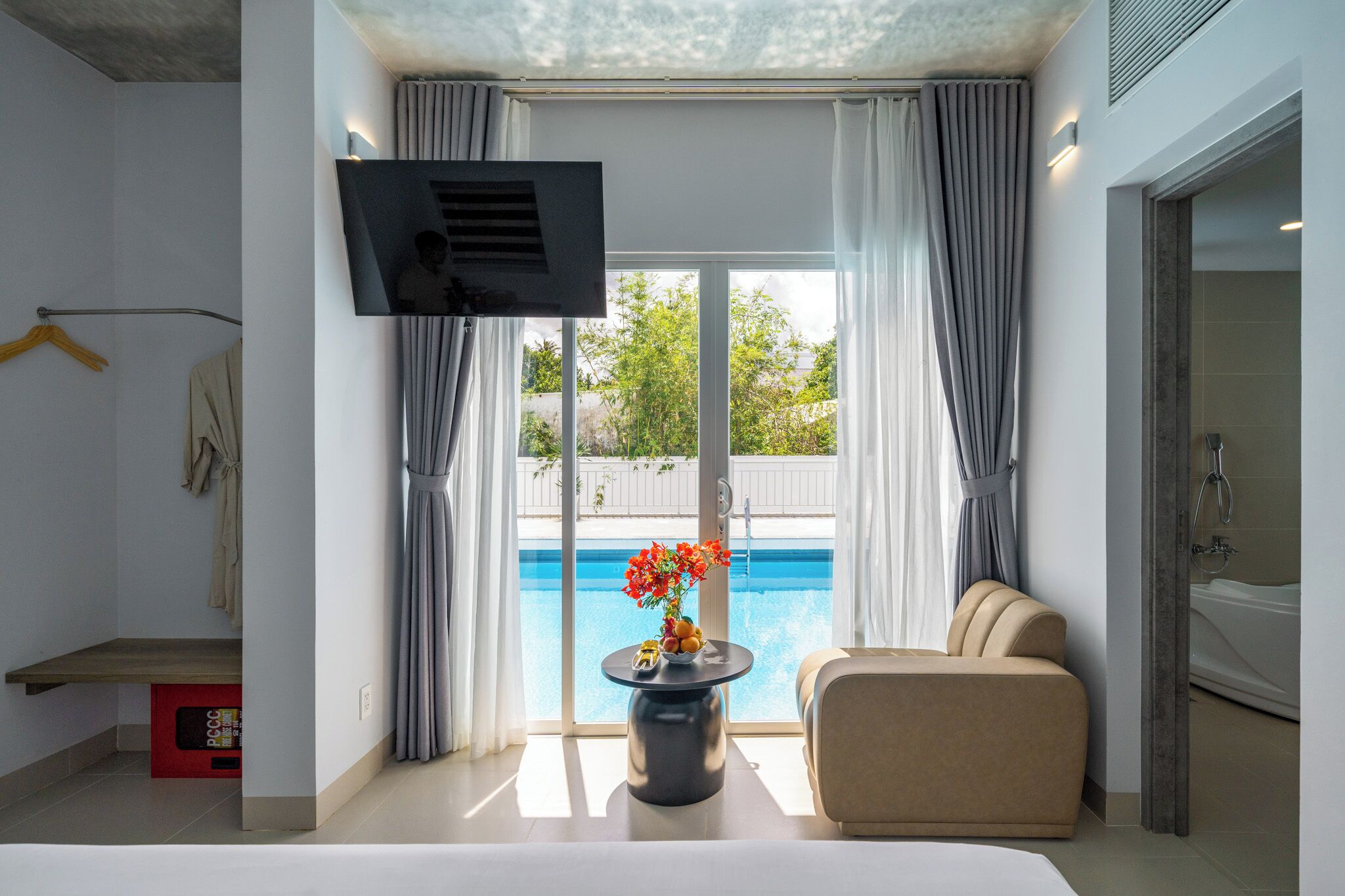 A modern hotel room with a large window, a sofa, a small table with a vase, and a TV mounted on the wall.