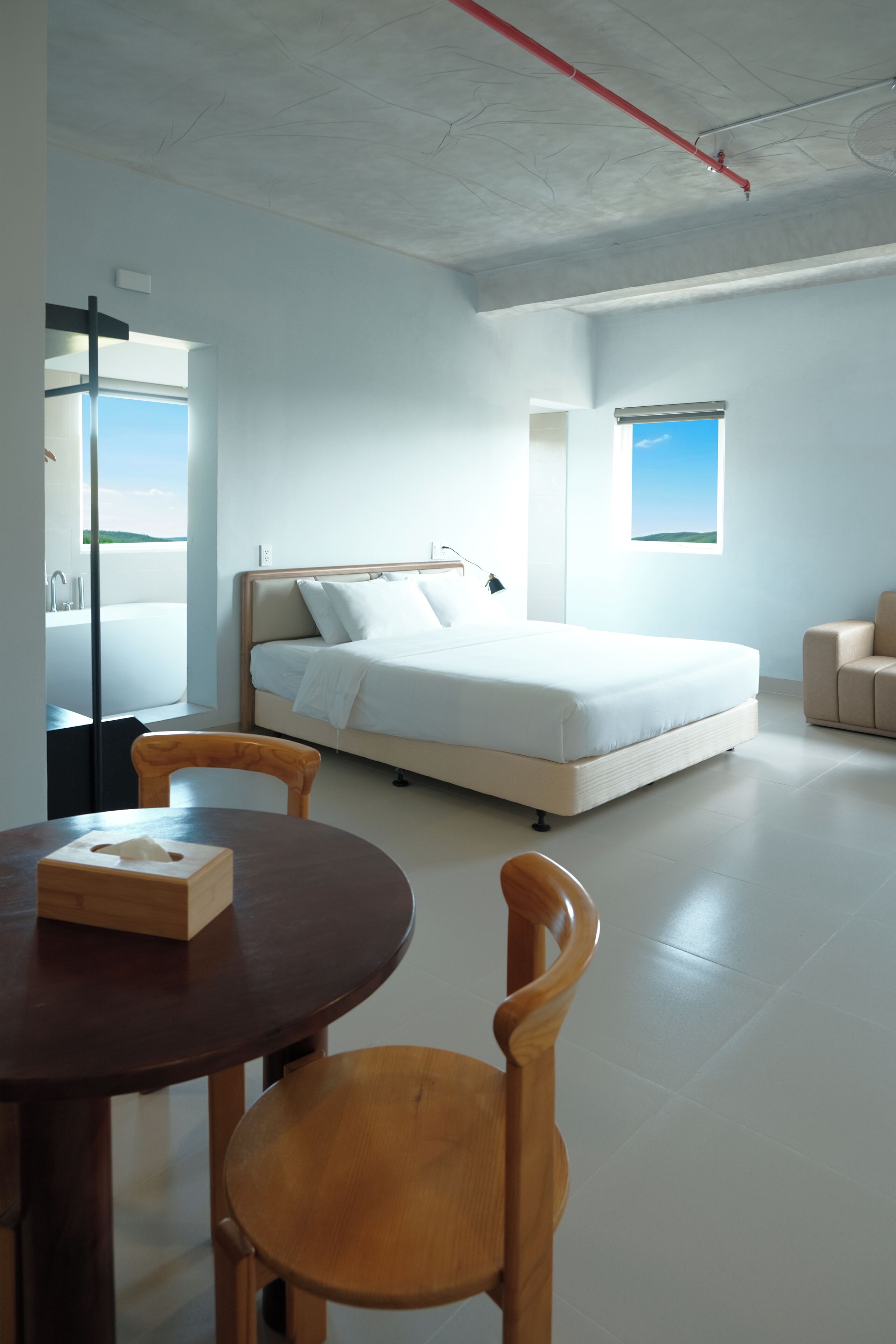 A modern hotel room with a large bed, a wooden dining table, and a view of the sky through a window.