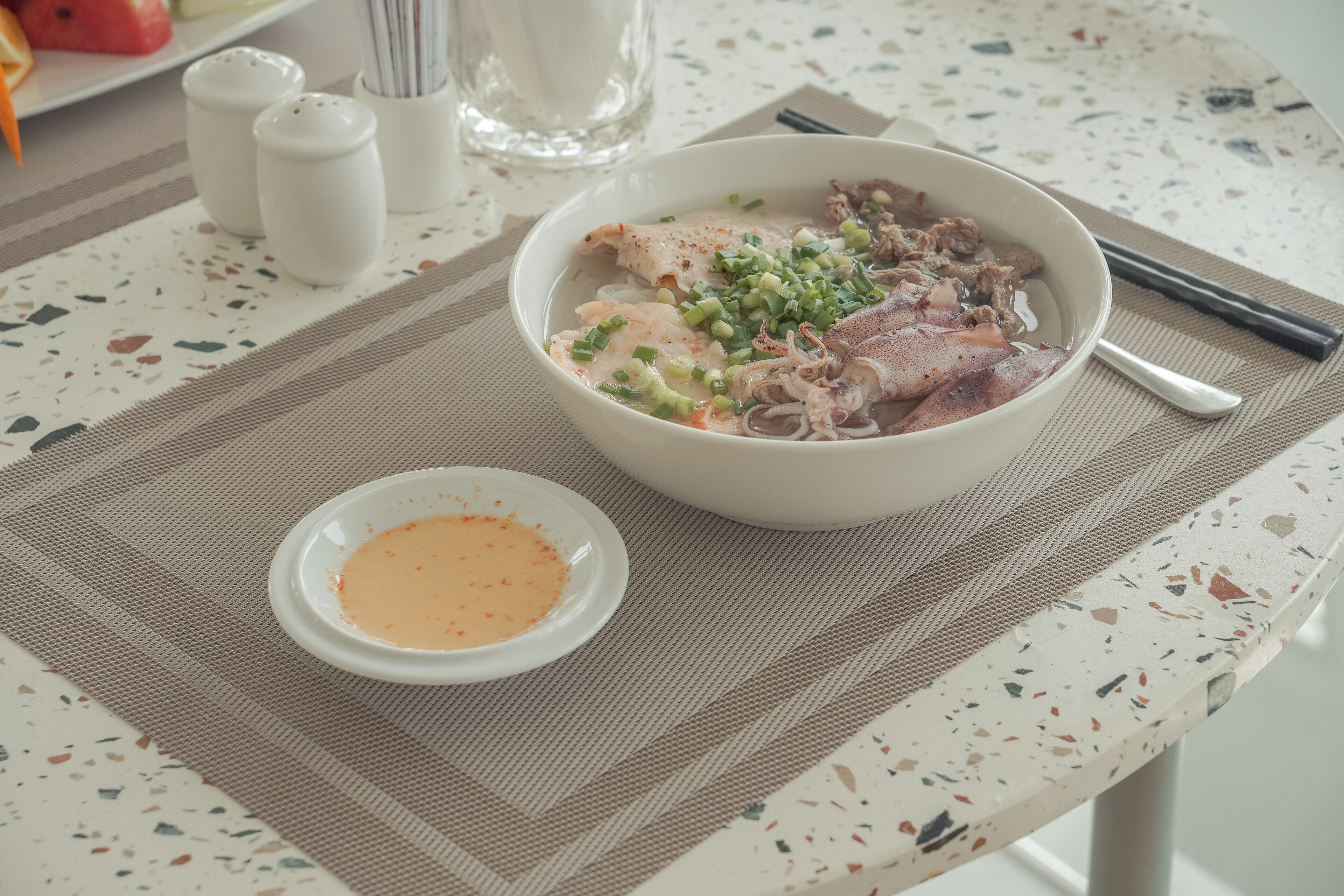 Eine Schüssel Suppe mit Stäbchen und einer kleinen Soßen-Beilage auf einem Tisch mit Unterlage.