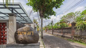 Monumento de piedra con el nombre 'Dewari' y una placa con texto, situado al lado de un moderno edificio con techo de vidrio, junto a un camino pavimentado.